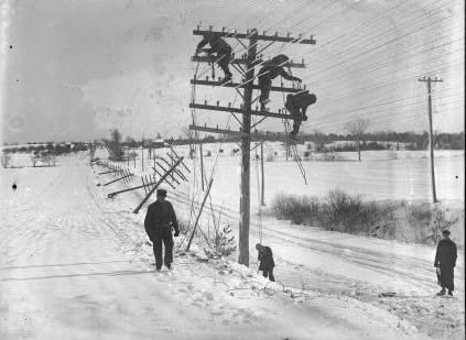 The photograph shows a winter line-repair scene in black and white. Three linemen work at the top of a utility pole strung with many wires, a row of damaged poles leans away down the left side of the snow-covered road with wires fallen toward the ground, a man in a long coat stands on the road in the foreground, and two smaller figures stand near the pole base and at the right edge. Snow covers the fields to the horizon.
