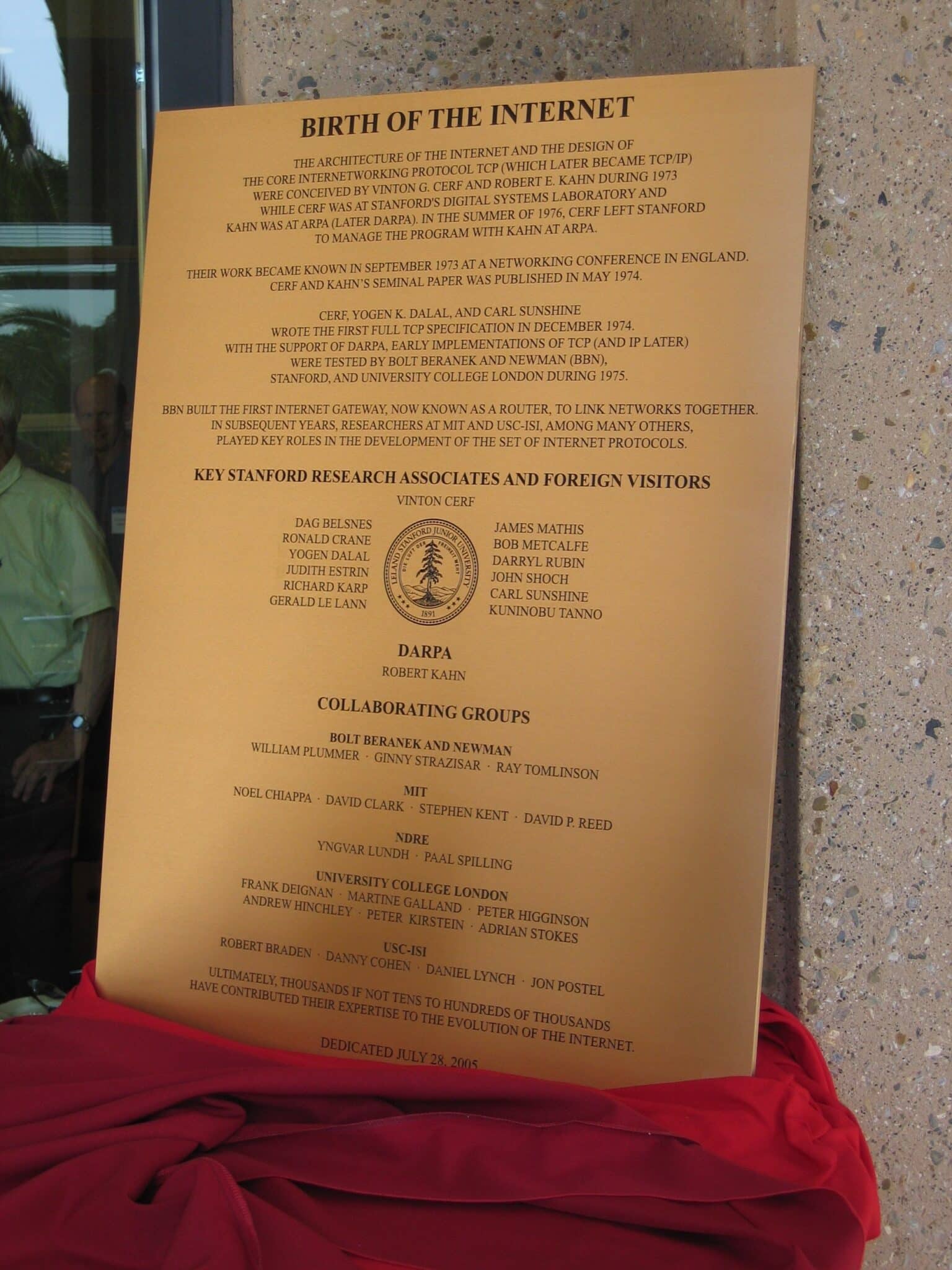 The photograph shows the Stanford University Birth of the Internet plaque at its unveiling, gold toned, leaning against a stone pillar with the red ceremonial drape gathered at the base and attendees reflected in the glass door at the left edge. The cast text credits Vinton G. Cerf and Robert E. Kahn with conceiving the architecture of the Internet and the design of the protocol TCP during 1973, a roster of thirty-three names surrounds the Leland Stanford Junior University seal, and the cast dedication date reads July 28, 2005.